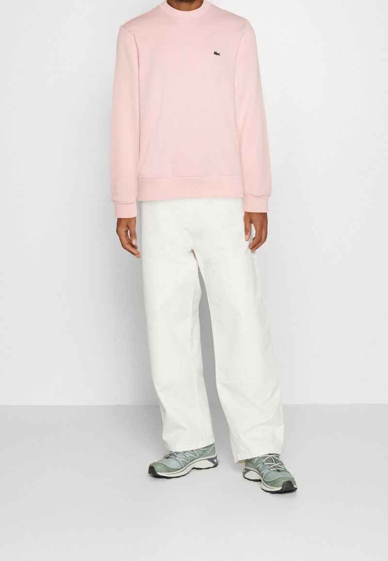 Personne portant un sweat-shirt à manches longues rose pâle, un pantalon blanc à jambes larges et des chaussures de sport vertes, debout devant un fond clair uni.
