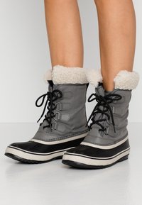 Bottes de neige grises avec une tige texturée, un bord en fausse fourrure beige, une pointe et une semelle en caoutchouc noir, et des rayures blanches contrastantes. Design à lacets.