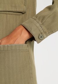 Tkanina olivově zelené barvy s vzorem herringbone, s manžetou a zapínáním na dva knoflíky. Obsahuje boční kapsu a měkký, texturovaný povrch.