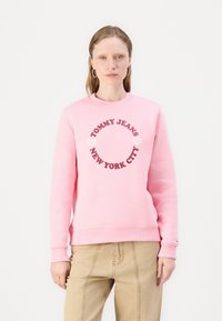 Lys rosa sweatshirt med rundt logo med teksten "TOMMY JEANS" og "NEW YORK CITY" i mørk rosa, myk bomullsstoff, ribbede mansjetter og nederkant.