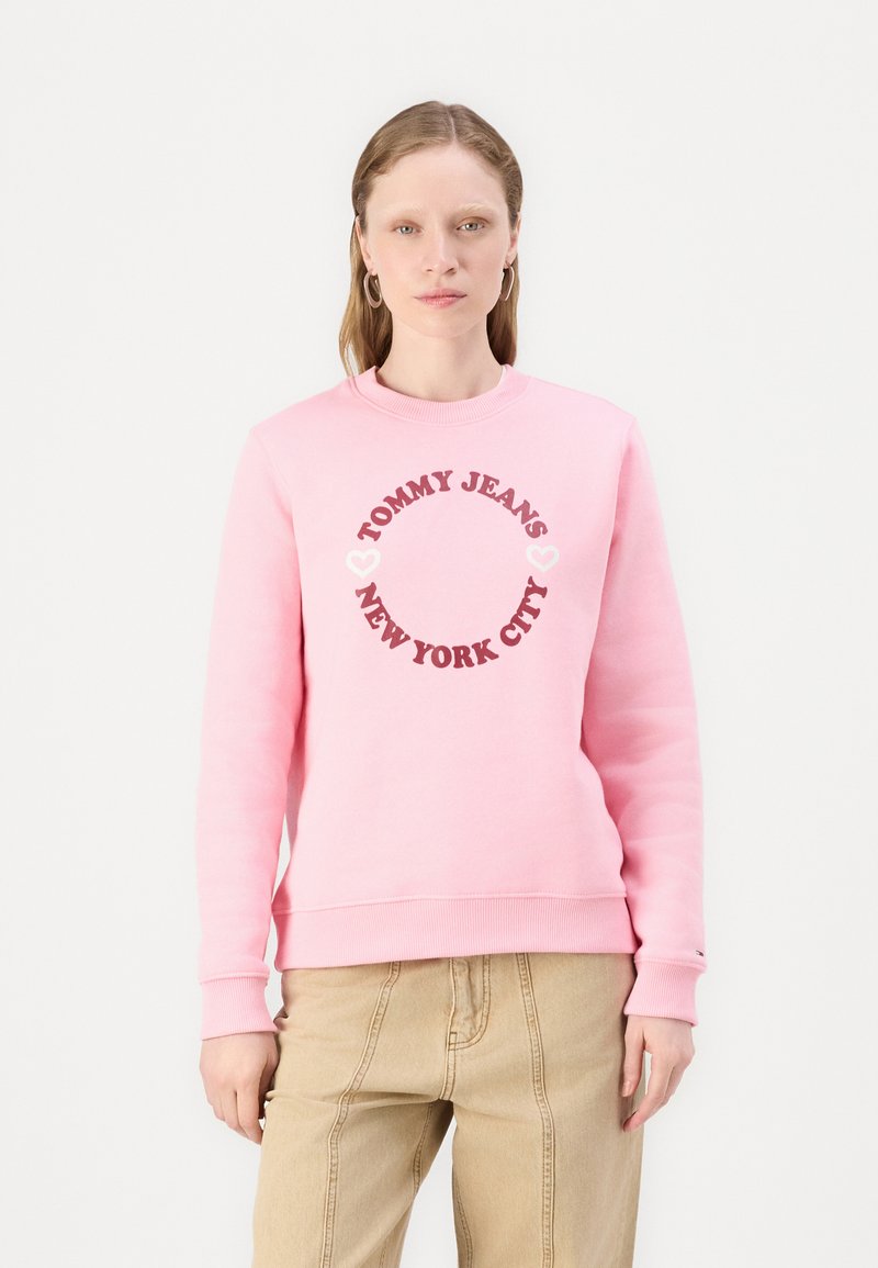 Lys rosa sweatshirt med rundt logo med teksten "TOMMY JEANS" og "NEW YORK CITY" i mørk rosa, myk bomullsstoff, ribbede mansjetter og nederkant.