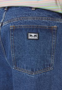 Vaqueros de mezclilla azul que presentan un bolsillo trasero con un parche de logo en blanco y negro, costuras naranjas visibles y un acabado de tela texturizada.