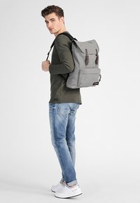 Zaino in tessuto grigio con chiusura a pattina, dotato di spallacci in pelle marrone e una tasca frontale, indossato da una persona in abbigliamento casual.