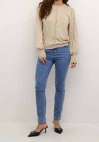 Béžový svetr na knoflíky s dlouhým rukávem, spárovaný s světle modrými úzkými džínami a černými lodičkami s mašlí na špičce.