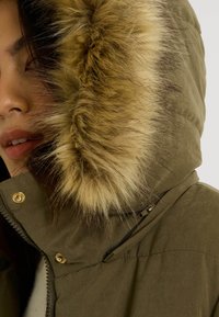 Parka verde aceituna con capucha forrada de piel. Cuenta con herrajes dorados y cierre frontal con cremallera. Tela lisa con una apariencia ligeramente textura.
