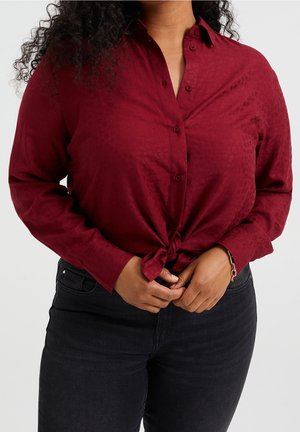 Person trägt ein langärmliges, burgunderfarbenes Hemd mit Knöpfen, das an der Taille gebunden ist, dazu schwarze Jeans, die Hände sind vor dem Körper verschränkt.