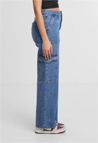 Pantalones cargo de pierna ancha en denim azul claro, con bolsillos laterales y traseros, y cierre de cremallera y botón. Complementados con zapatillas casuales.