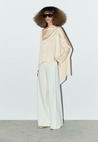 Top in satin beige chiaro con maniche ampie, abbinato a pantaloni bianchi a gamba larga e scarpe dorate metallizzate. Occhiali da sole scuri oversize.