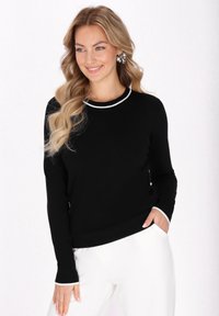 Un pulover negru cu mâneci lungi, având un guler alb cu nervuri. Materialul pare neted; modelul poartă pantaloni albi și are o croială lejeră.