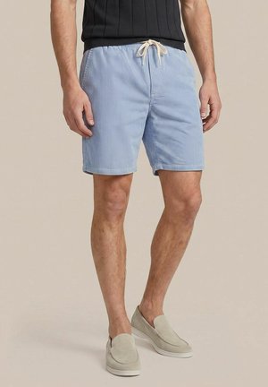 Man draagt lichtblauwe gestreepte korte broek met een witte trekkoord, beige instapschoenen en een zwart geribd mouwloos topje.