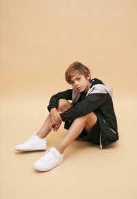 Svart och grå jacka med huva, svarta shorts, sittande mot en ljus orange bakgrund, iförd vita sneakers med perforerad detalj.