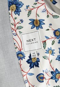 Fodera floreale con fiori blu e oro su uno sfondo bianco; tessuto grigio a sinistra; include un'etichetta bianca con la scritta "NEXT TAILORING."
