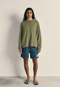 Langærmet olivengrøn skjorte med et lille rødt logo, parret med denimshorts og flip-flops, stående på en neutral baggrund.
