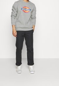 Persona vistiendo un sudadera gris de Dickies, pantalones negros remangados y zapatillas blancas, de pie frente a un fondo blanco liso.