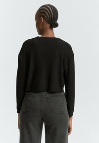 Schwarzer, kurzer Pullover mit langen Ärmeln, aus strukturiertem Strickstoff. Kombiniert mit hochtaillierten grauen Jeans aus Denim mit Gesäßtaschen.