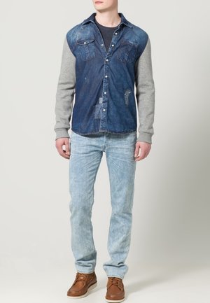 Spijkershirt met grijze mouwen, lichtgewassen jeans en bruine schoenen. Het shirt heeft knopen, zakken en verweerde accenten.