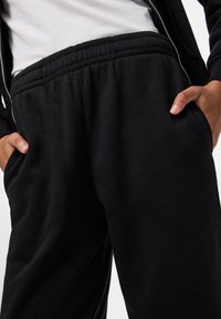 Juodos sportinės kelnės su elastinga juosmeniu, sklandžiu paviršiumi ir šoniniais kišeniais. Medžiaga atrodo minkšta ir stora, tinkama kasdieniam dėvėjimui.