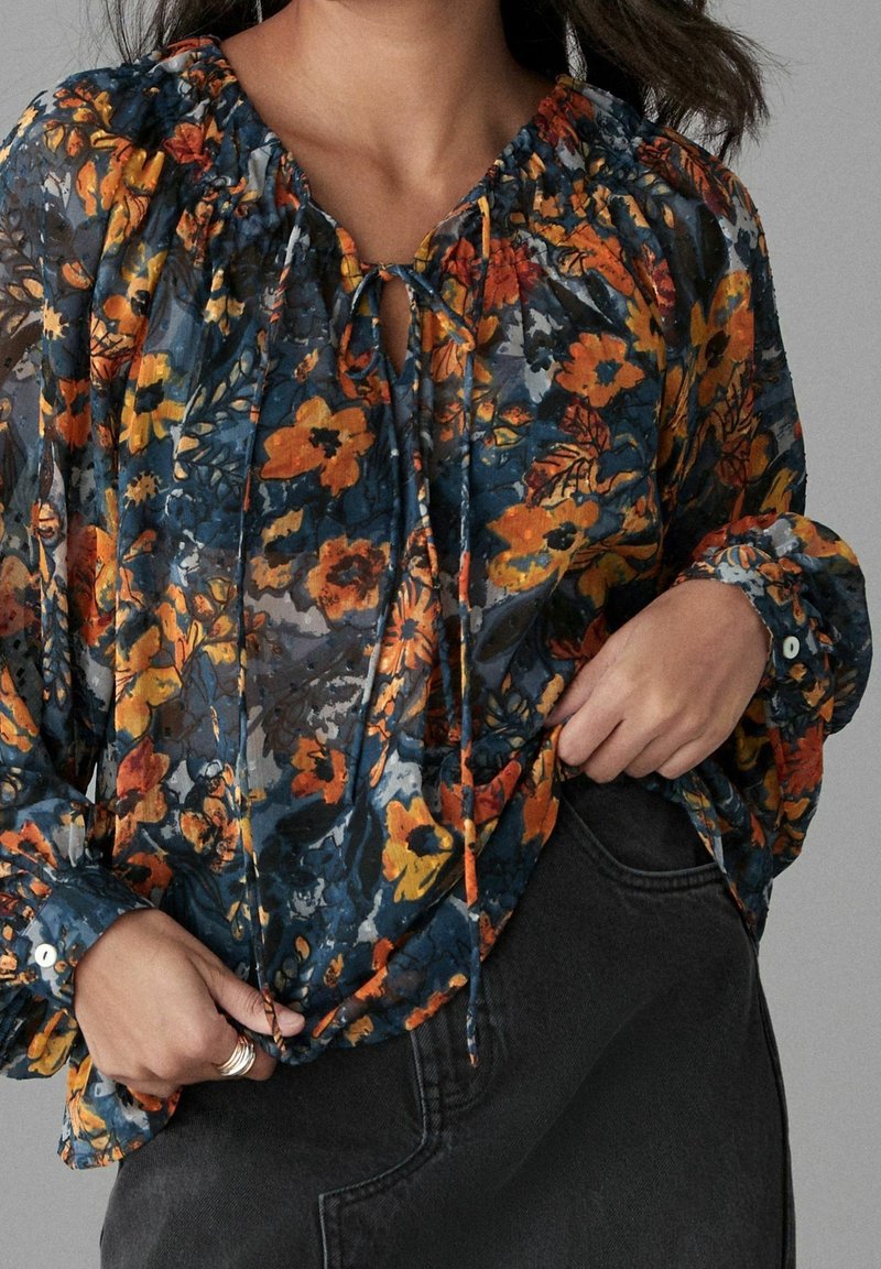 Blouse fluide à fleurs en tissu transparent, avec une base bleu foncé ornée de fleurs orange et jaunes, un col froncé et des poignets boutonnés.