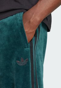 Mano de piel oscura en el bolsillo de unos pantalones de terciopelo verde azulado con logo y rayas negras de Adidas, combinados con una camiseta negra.