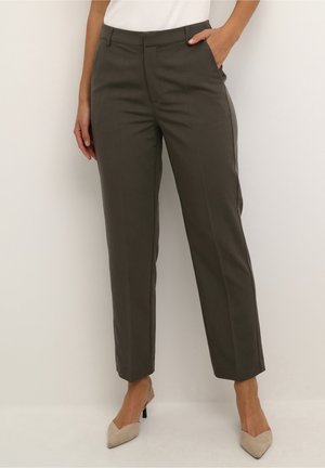 Kobieta w ciemnooliwkowych, dopasowanych spodniach z nogawkami do kostek, z jedną ręką w kieszeni, w beżowych, zgrabnych butach na niskim obcasie, na jednolitym tle.