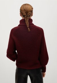 Burgundin neulepusero korkealla kauluksella, jossa on teksturoitu muotoilu ja pallohiha. Takana näkyy letitetty kampaus.