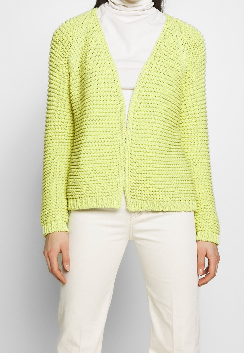 Dzianinowy kardigan w jasnym limonkowym kolorze, o luźnym kroju, z otwartym przodem, ściągaczami na mankietach i teksturowanym przeszyciem. Noszony na białym golfie.