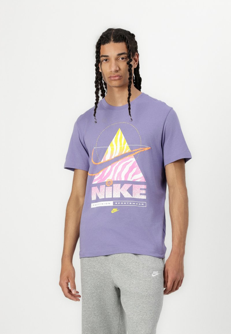 Purpura T-krekls ar grafisku dizainu, kas attēlo dzeltenu un rozā piramīdu, tekstu "NIKE" un apaļu akcentu. Savienots ar pelēkiem sporta biksēm.