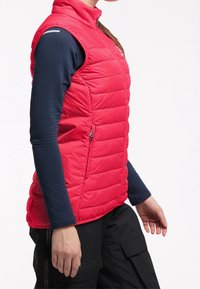 Henkilö, joka on pukeutunut punaisiin tilkkuhameisiin ilman hihoja merivoimien pitkähihaisen paidan ja mustien cargo-housujen päälle, poseeraa sivuprofiilissa valkoista taustaa vasten.