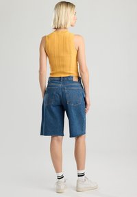 Keltainen ribbed ilman hihoja oleva toppi yhdistettynä sinisiin farkkushortseihin, joissa on raaka helma. Mallilla on päällä valkoiset tennarit mustilla raidoilla olevien sukkien kanssa.