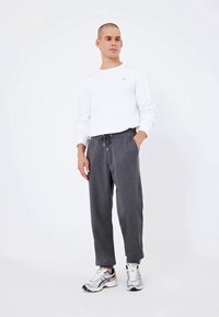 Weißes Langarmhemd mit kleinem goldenen Logo, kombiniert mit grauen Jogginghosen mit elastischem Bund und seitlichen Taschen. getragen mit Sneakers.