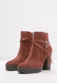 Stivali ankle in suede marrone con tacco rialzato, cinturino in pelle decorativo, chiusura con zip laterale e suola in gomma texturizzata.