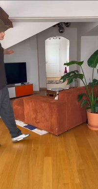 Oranžinė velveto dviejų dalių sofa kvadrato formos, stovinti ant medinio grindų paviršiaus, šalia žaliosios augalijos ir rudos apvalios stalo.