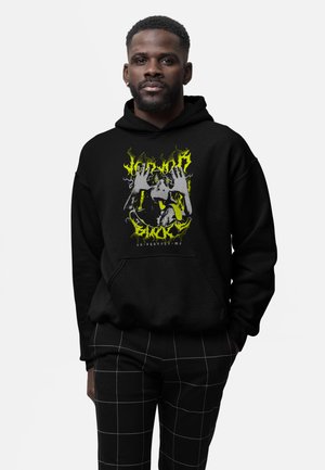 Homme portant un sweat à capuche noir avec un visuel jaune représentant des mains de singe et un texte abstrait, associé à un pantalon noir avec des lignes de grille blanches.
