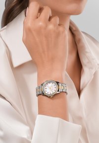 Sudraba un zelta toņu rokas pulkstenis ar apaļu baltu ciparnīcu, kristāla akcenti un metāla siksna, kas veidota no pārmaiņus izvietotām saitēm.