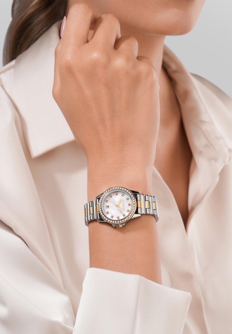 Sudraba un zelta toņu rokas pulkstenis ar apaļu baltu ciparnīcu, kristāla akcenti un metāla siksna, kas veidota no pārmaiņus izvietotām saitēm.