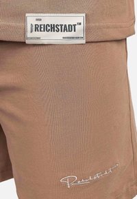 Pantaloni corti beige con un'etichetta bianca prominente con la scritta "REICHSTADT™" e cuciture decorative bianche. Texture liscia e design aderente.