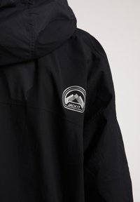 Sort vandtæt jakke med hætte, glat tekstur og et hvidt rundt patch med bjerge og ordet "JACKIT" på skulderen.