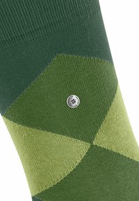 Primo piano di un calzino verde e verde chiaro con un motivo, caratterizzato da un piccolo logo argentato. La trama è visibile, mettendo in risalto il design del tessuto.