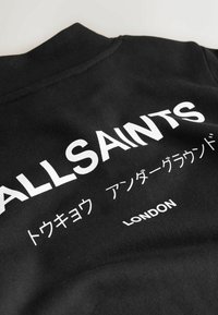 Melna sporta krekla ar apaļu kaklu, uz kura ir izcili balti burti "ALLSAINTS" un citi simboli vairākās valodās. Mīksta, teksturēta auduma.