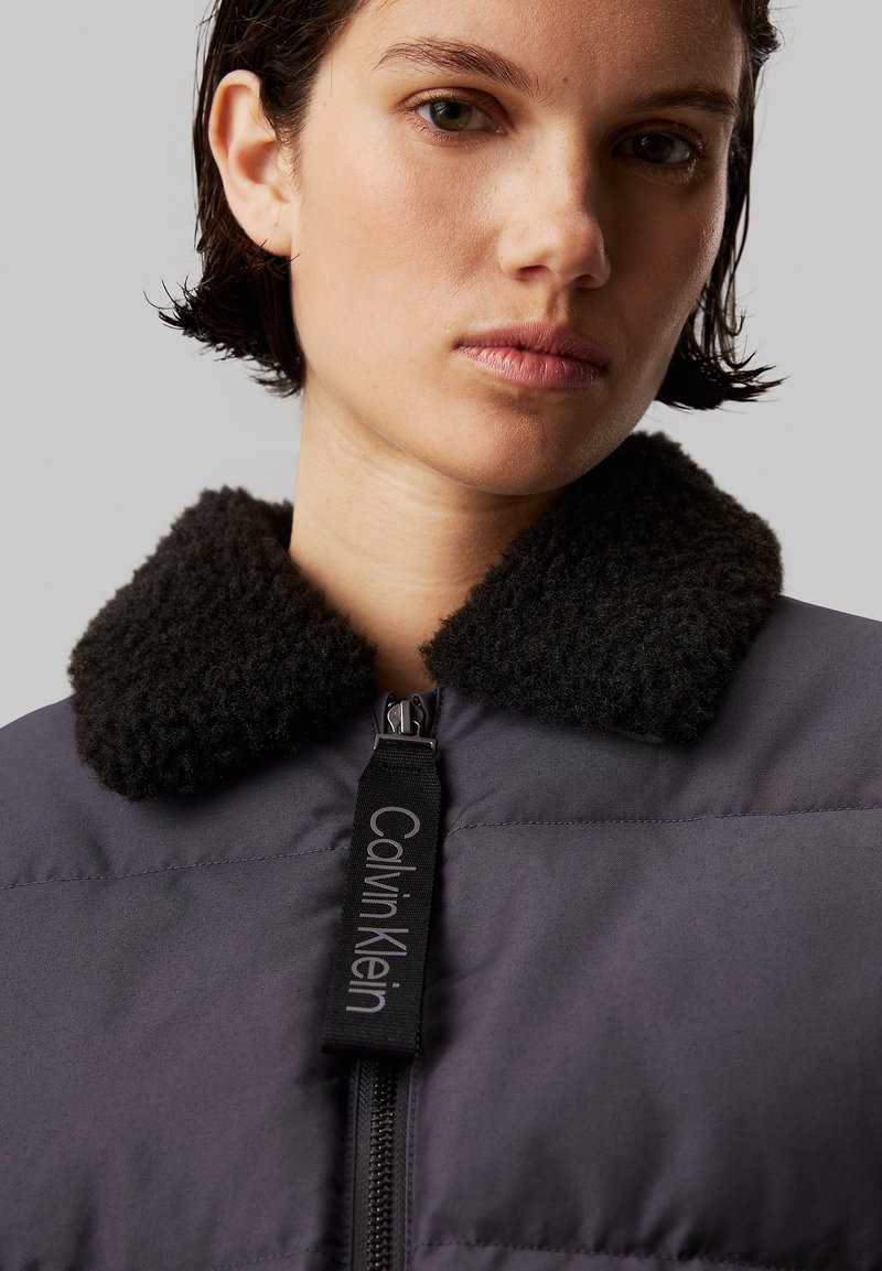 Giacca Calvin Klein con un corpo imbottito grigio scuro, colletto nero peloso e un'etichetta con logo. Presenta una zip frontale e una texture liscia.