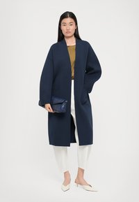 Manteau bleu marine à coupe décontractée, avec un col châle, une ouverture à l'avant et des poches latérales, associé à un pantalon blanc et une pochette texturée bleu marine.