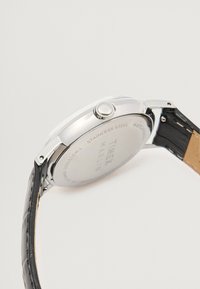 Caixa de relógio em aço inoxidável com um acabamento prateado polido, apresentando uma pulseira de couro preto texturizado e uma coroa tradicional para ajuste.