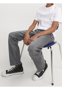 Persona vestida con una camiseta blanca, jeans grises y zapatillas altas negras sentada en una silla azul con patas de cromo, con las manos descansando sobre las piernas.