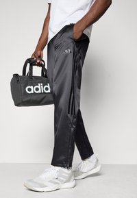 Sorte træningsbukser med sidelommer, tætsiddende design og lynlåslommer; hvid duffel taske med logo; lyse træningssko.