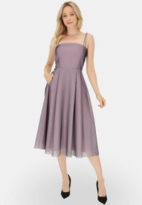 Fioletowa sukienka bez rękawów z prostym dekoltem, rozkloszowanym dołem, kieszeniami i przezroczystą nakładką; zaprezentowana w czarnych szpilkach na białym tle.
