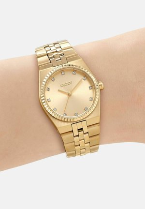 Goldfarbene DKNY-Armbanduhr mit Kristallstundenmarkierungen und verbundenem Metallband, getragen an einem hellhäutigen Handgelenk, zeigt 1:50 Uhr an.