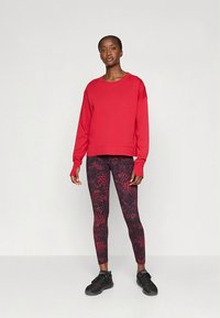 Piros pulóver hosszú ujjakkal és kerek nyakkivágással, fekete-piros mintás leggingsszel párosítva. Fekete sportcipővel viselve.