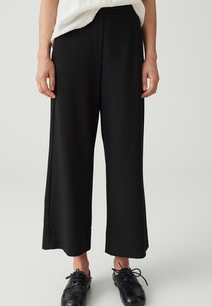 Person trägt lockere schwarze kurze Hosen und schwarze Schnürschuhe, steht vor einem einfarbigen hellen Hintergrund.