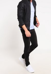 Giacca di pelle nera con spalle trapuntate, t-shirt azzurra chiara, jeans neri strappati e sneakers bianche. Focalizzazione su uno stile casual moderno.