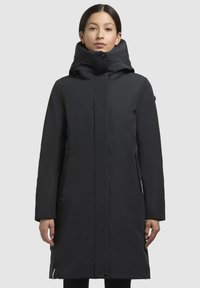 Manteau noir long avec un col haut et deux zips sur les côtés. Tissu lisse, coupe droite et design minimaliste sans motifs ni accents.